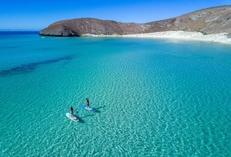 White sand beach view from above, with crystal-clear waters and a couple practicing paddle-boarding in La Paz, Mexico.