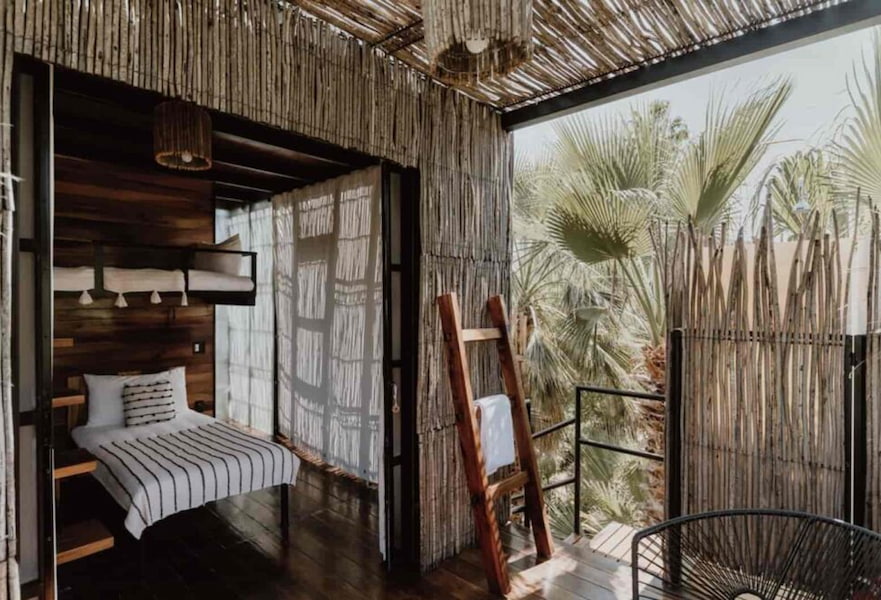 Treehouse interior with wooden floor and a white clean bed with palm trees outside.
