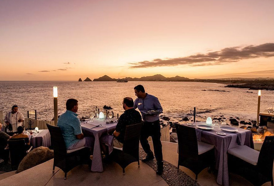 Two men seated at a table and getting served by a waiter in one of the best Cabo San Lucas restaurants with an ocean view.