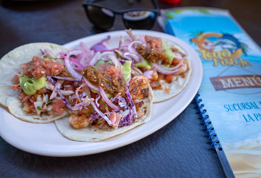 Three delicious shrimp tacos with corn tortillas and Mexican salsas on top with Claro Fish Jr. menu on the side, La Paz, Mexico.