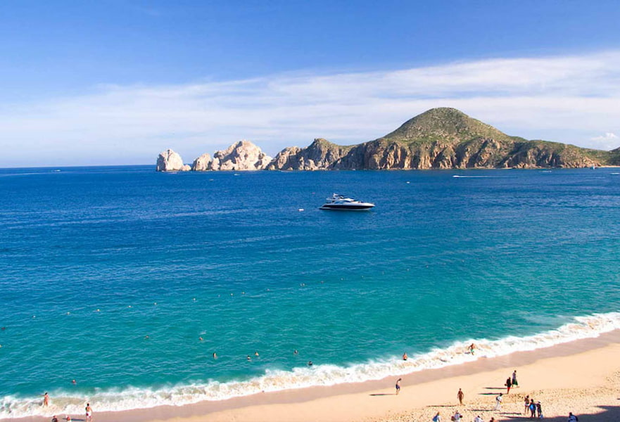 Luxurious yacht sailing in the ocean with The Arc mountains behind and Medano beach with few people walking on the seashore, Cabo San Lucas, Mexico.