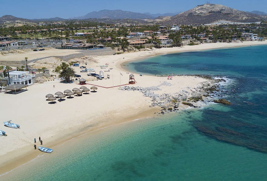 Palmilla beach in Mexico with palapas, boats, a lifeguard kiosk, and resorts with palm trees on the distance.