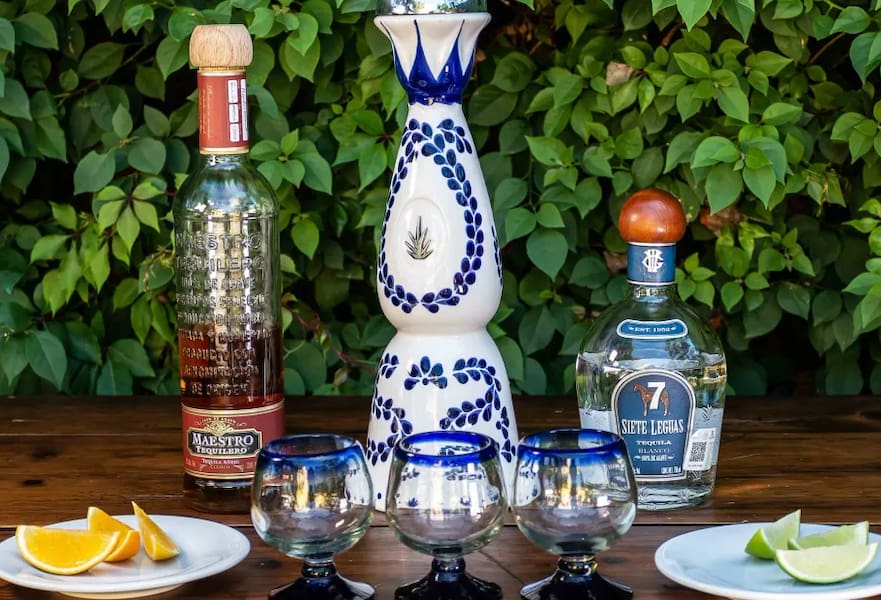 Three tequila bottles facing three empty glasses in a wooden table with orange and lime slices on the sides