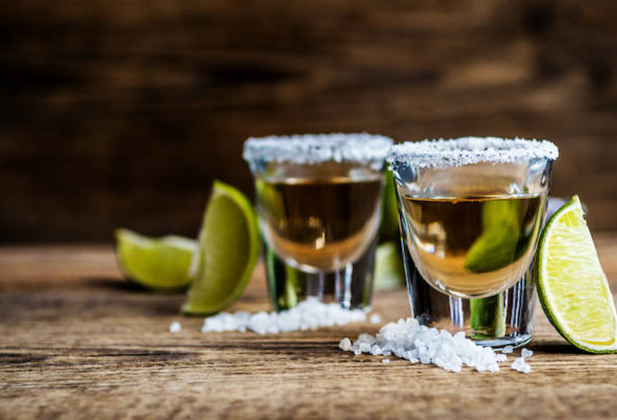 Two tequila shots served in salted glasses and lime slices on a wooden surface