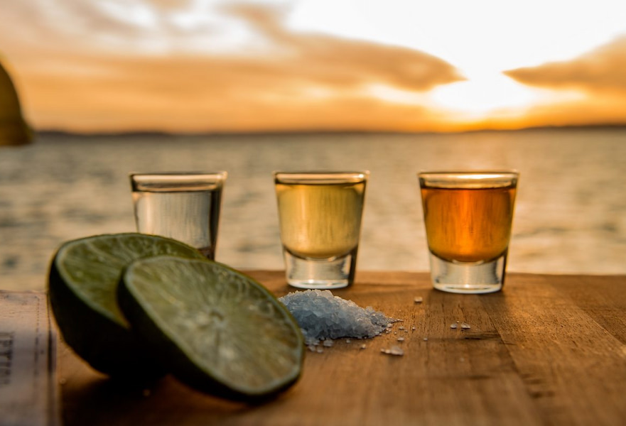 Three tequila shots with salt and two lemon slices in a wooden table with an ocean sunset backdrop.
