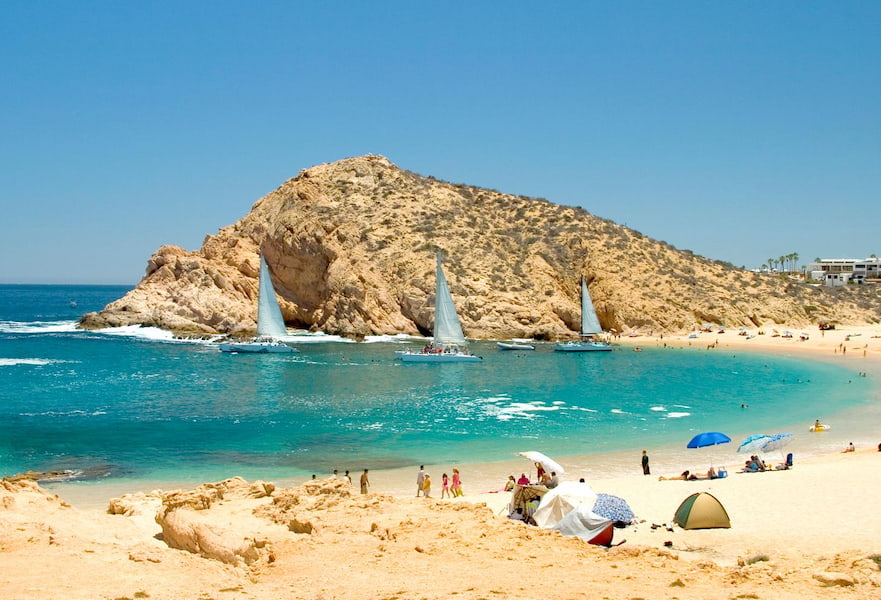 Cabo San Lucas coastal area, white sand with turquoise waters, people resting with shades and sailboats on the seashore.